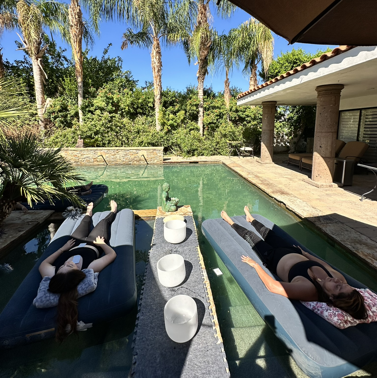 Women floating on pool rafts with crystal singing bowls surrounded by palm trees in an outdoor pool setting